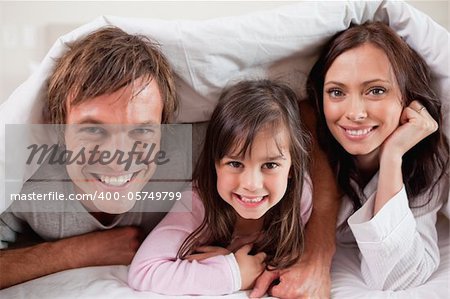 Happy parents lying under a duvet with their daughter in their bedroom