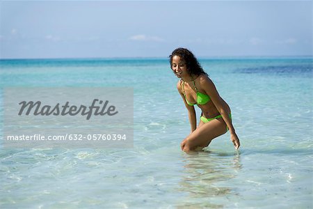 Young woman in bikini standing in ocean, bending to touch water, portrait