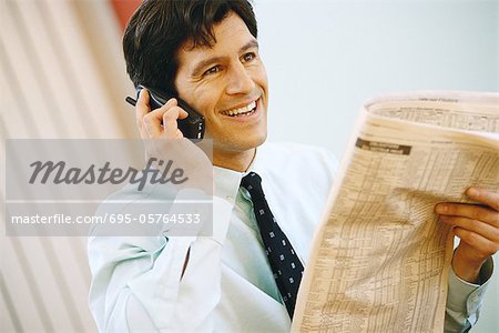 Businessman using phone and holding up financial section of newspaper, smiling