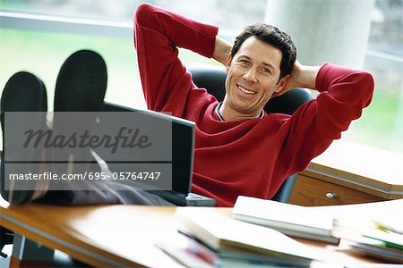 Mann, sitzend am Schreibtisch mit Füßen, Hände Laptop auf dem Schoß, halten hinter dem Kopf