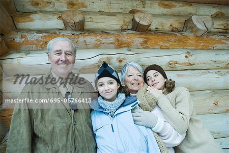 Senior couple standing with teen and preteen granddaughters in front of log cabin, smiling, portrait