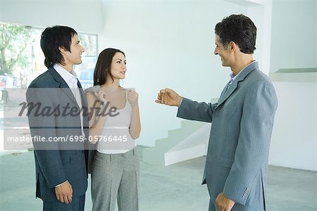 Real estate agent holding out keys to young couple, standing in bare home interior