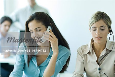 Two female colleagues, one using cell phone, the other using headset