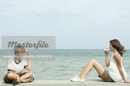 Boy and teenage girl sitting near ocean, talking through tin can phone