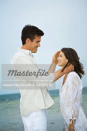 Man standing face to face with young female companion on beach, pushing her hair back, side view
