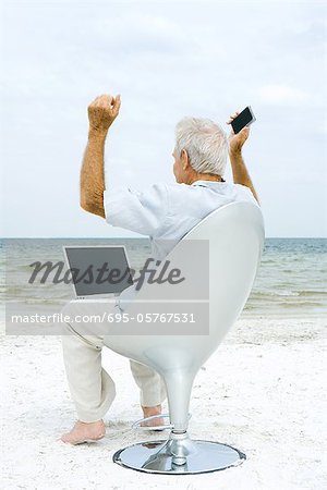 Alter Mann mit Laptop und Handy am Strand, Anheben der Arme