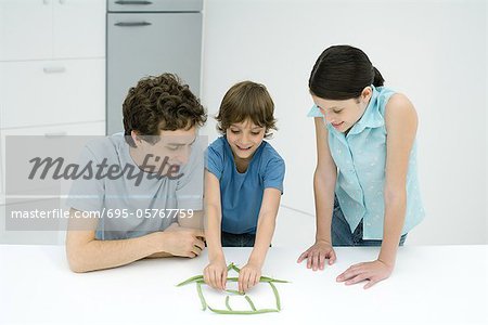 Little boy in kitchen with father and sister, building house out of green beans, all smiling