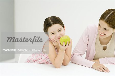 Kleines Mädchen sitzt am Tisch mit Mutter, Hand unter Kinn, Apfel, lächelnd in die Kamera halten