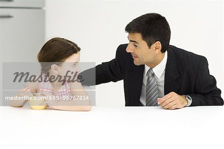Father and daughter sitting at table, looking at each other, little girl eating cereal