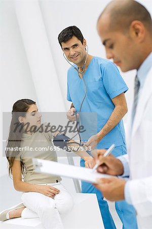 Male nurse measuring young patient's blood pressure, smiling at camera, doctor writing on clipboard in foreground