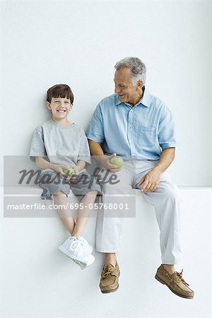 Grandfather and grandson sitting side by side on ledge, holding apples, both smiling