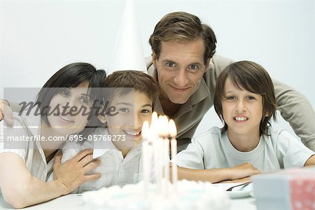 Family in front of birthday cake with lit candles, one boy wearing party hat