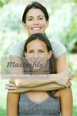 Mother standing behind teenage daughter, embracing her, both smiling at camera, portrait