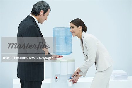 Two professionals chatting beside water cooler, woman filling disposable cup