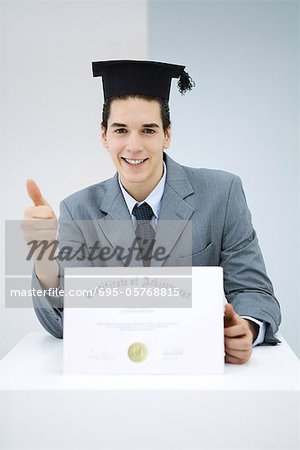 Young man with diploma, wearing graduation cap, making thumbs-up gesture