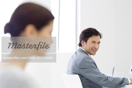 Professionelle Mann sitzt am Schreibtisch, lächelnd über die Schulter in die Kamera, die Kollegin im Vordergrund