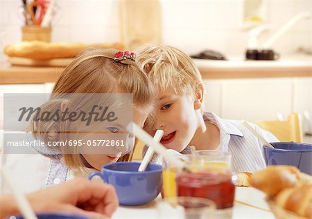 Children sitting at table leaning heads together, head and shoulders, cups, jelly jar and adult hand blurred in foreground