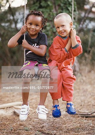 Petit garçon et une fille assis swing ensemble, chacun tenant un côté de la corde de la balançoire, souriant à la caméra, de pleine longueur.