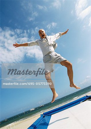 Homme mûr, sauter sur le trampoline sur la plage, les bras tendus, la bouche ouverte