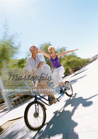 Mature couple riding together on tandem bike, woman holding her arms out, blurred