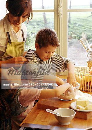 Two children and mother cooking in kitchen, children with hands in bowl, mother in apron smiling