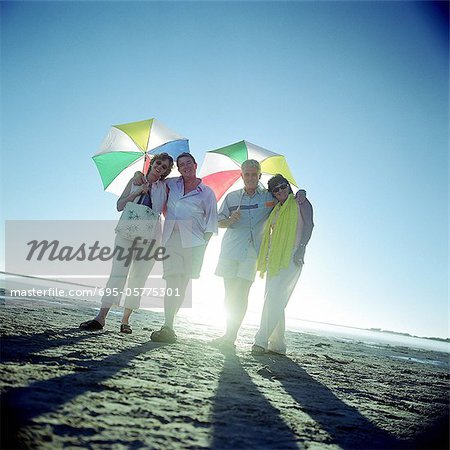 Two mature couples standing on beach, holding umbrellas