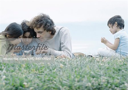 Man lying on grass with boy and girl looking at cellphone, second boy sitting apart