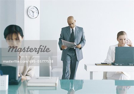 Male executive standing in office reading newspaper, female colleagues working on laptop computers