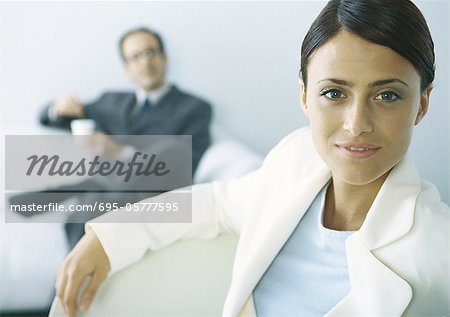Businesswoman looking at camera, businessman with legs crosses holding cup in background