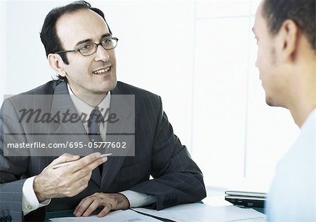 Businessman gesturing with pen in hand, speaking to man