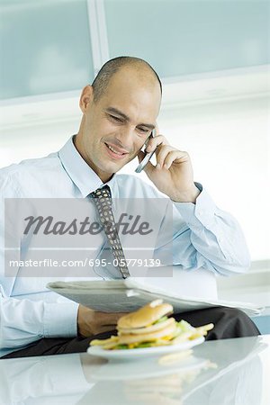 Man using phone and reading newspaper, hamburger on table in foreground