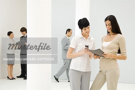 Two businesswomen standing together, looking down at folder, one holding cell phone, coworkers in background