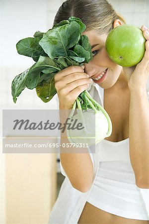 Woman holding up and hiding behind fresh kohlrabi and apple, smiling at camera
