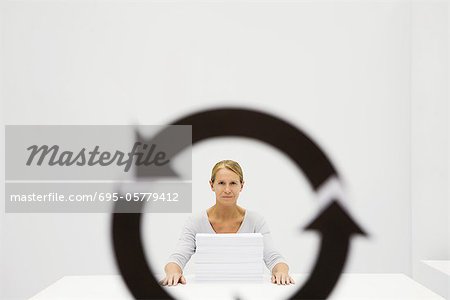 Professional woman sitting behind stack of paper, looking at camera through recycling symbol