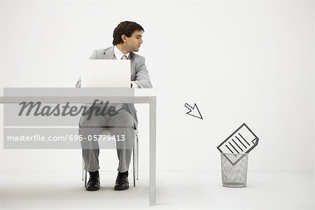 Businessman sitting at desk, looking at computer cursor pointing to document in trash can