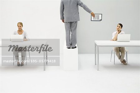 Professional women working with laptops, man standing between them, handing document to one