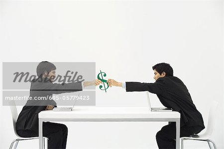 Businessmen sitting face to face at table, each reaching to hold dollar sign