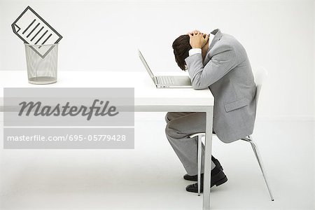 Businessman sitting at desk with head down, document in trash can nearby