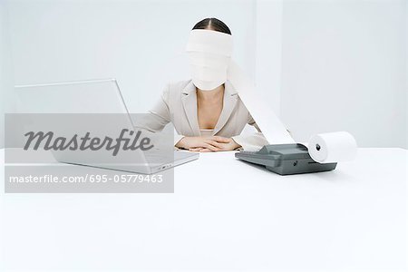 Woman sitting at desk with adding machine and laptop, printout tape wrapped around head