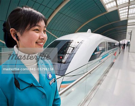 A smiling female attendant wearing a mobile phone earplug standing next to a Magnetic Levitation train on railway platform in Shanghai, China, Asia