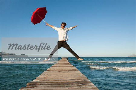 Jeune homme sautant sur la jetée avec parapluie Jeune homme sautant sur la jetée avec parapluie