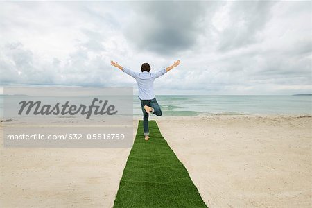 Man standing on one leg at end of carpet on beach with arms outstretched, rear view