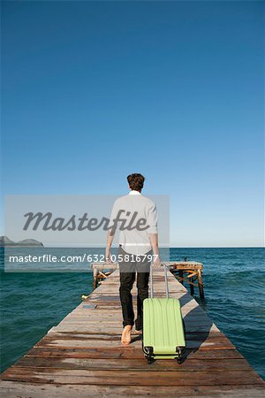 Man pulling luggage walking towards end of pier, rear view