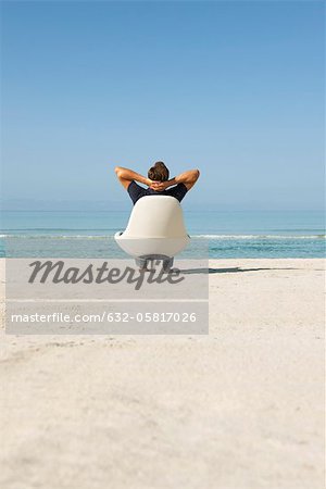 Man sitting in armchair on beach looking at ocean, rear view