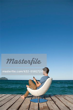 Young man sitting in armchair by lake using cell phone, side view