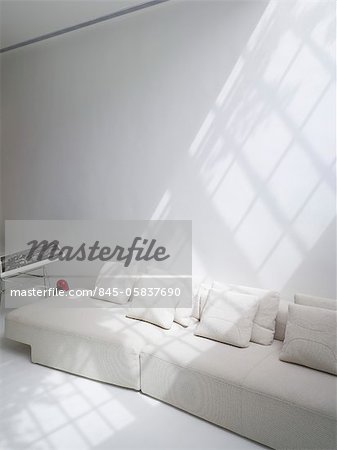 Moderne salon blanche avec fenêtre de haut niveau réparties entre de multiples barres de vitrage, blanc canapé avec des coussins et soleil en streaming pour les ombres, la lumière du soleil. Architectes : Hilit