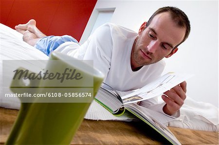 Homme couché dans son lit, lisant un livre, mug en avant-plan