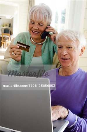 Senior man using laptop computer, senior woman phoning, holding credit card