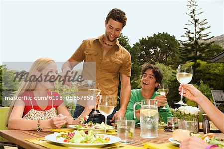 2 young smiling couples sitting at garden table, man pouring wine