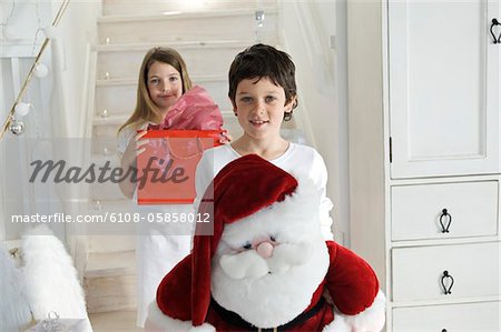 Christmas day, little boy holding a cuddly toy (Santa Claus), looking at the camera, sister with present in background, indoors